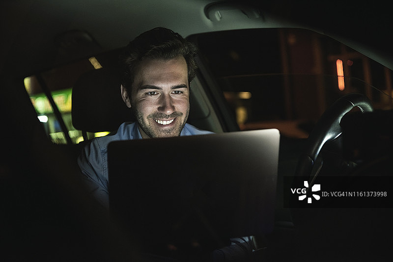 微笑的年轻男人晚上在车里使用笔记本电脑图片素材