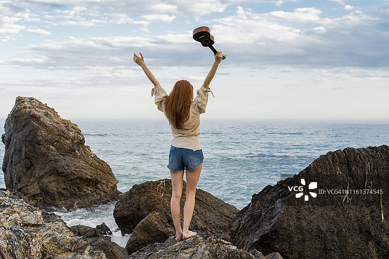 年轻女子站在岩石上，手拿尤克里里琴，眺望大海的背影图片素材