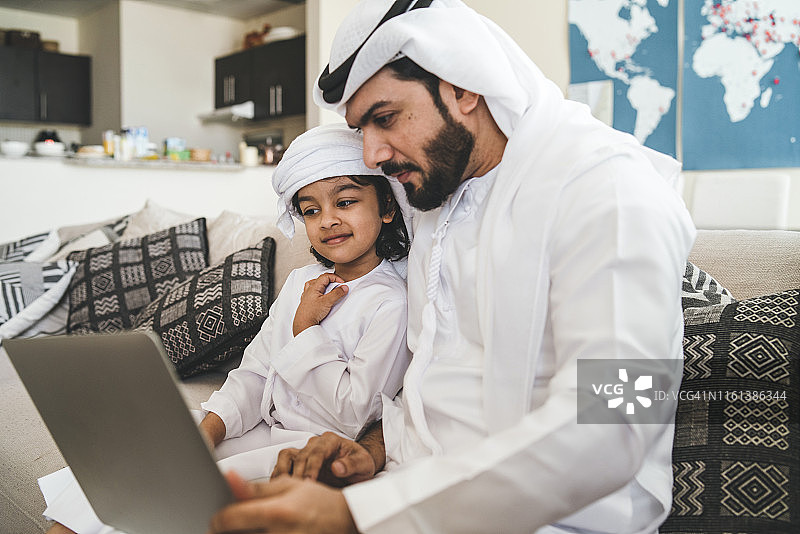 阿拉伯男人和儿子在家里的沙发上使用笔记本电脑图片素材