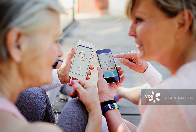 年长的女性朋友跑步者拿着智能手机坐在户外聊天图片素材