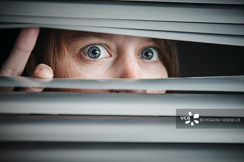 年轻女子睁大眼睛透过百叶窗向外看图片素材