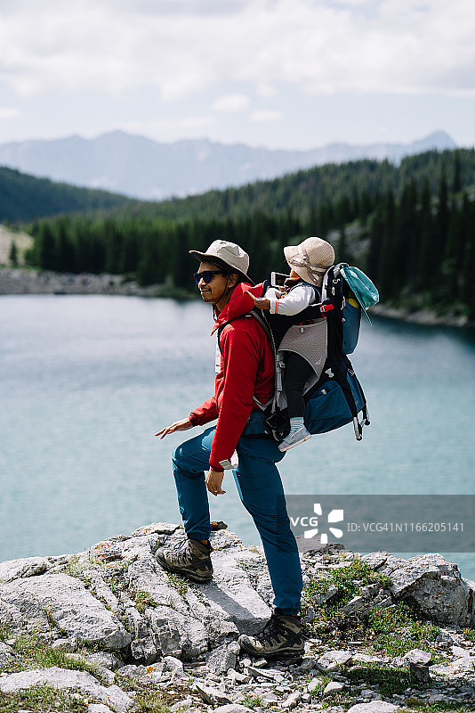 徒步男子背着幼儿站在湖泊和森林前的山中图片素材