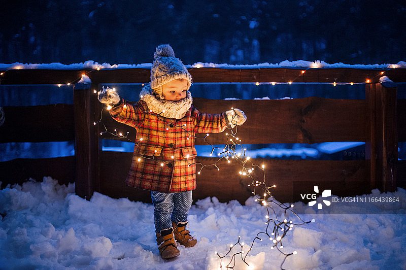 在冬天的户外，手持灯链站在露台上看雪的小女孩图片素材