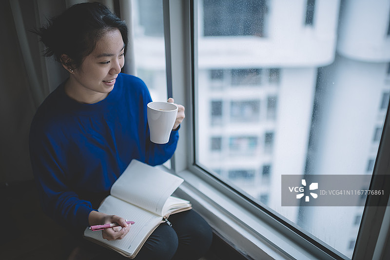 中国亚洲女学生坐在卧室的床边，拿着咖啡杯，在笔记本上写字，露出牙齿的微笑图片素材