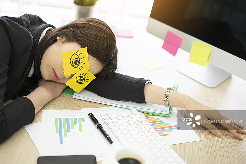 疲惫的女人在办公室的办公桌上睡觉，她在电脑旁画了假眼睛图片素材