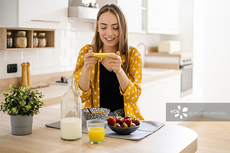 快乐的年轻女人在家中厨房使用手机并享用早餐图片素材