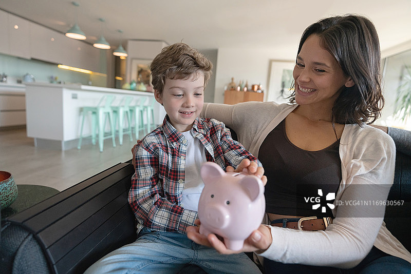 快乐的母亲和儿子在家坐在沙发上往储蓄罐里存钱图片素材