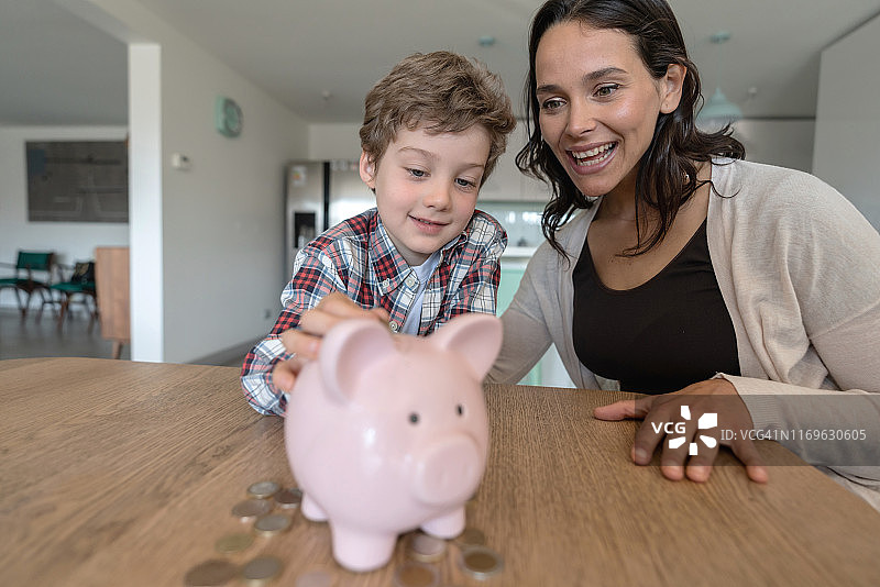 往存钱罐里投硬币的可爱小男孩和一旁兴奋微笑的妈妈图片素材