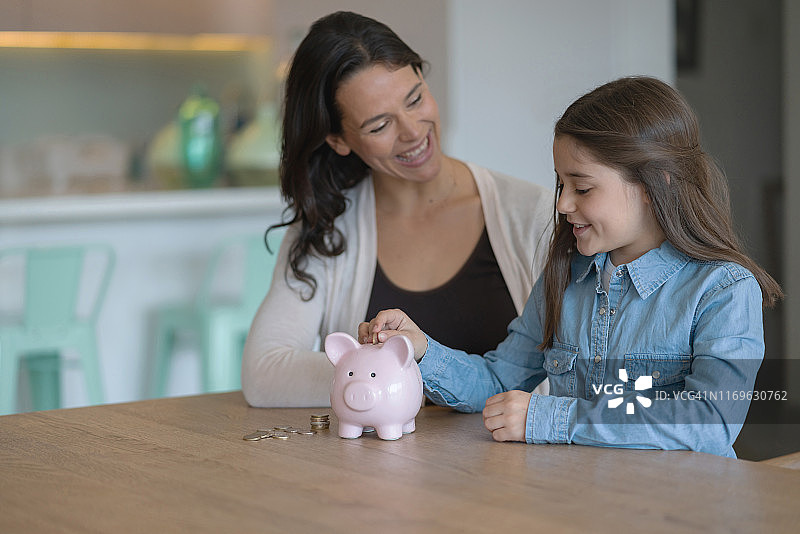 快乐的母亲坐在女儿旁边，看着她把硬币放进家里的存钱罐图片素材
