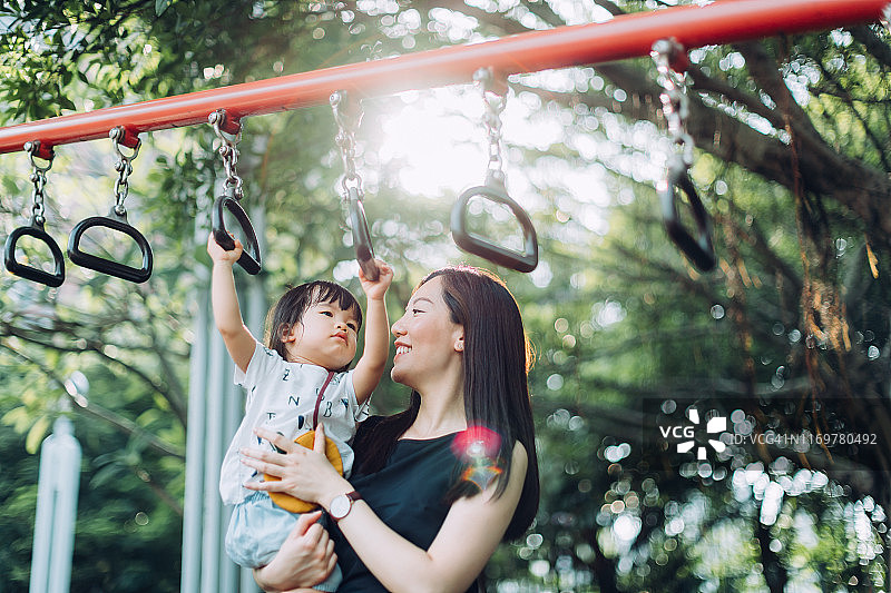 在阳光明媚的日子里，年轻的亚洲母亲在户外游乐场扶着女儿玩单杠图片素材