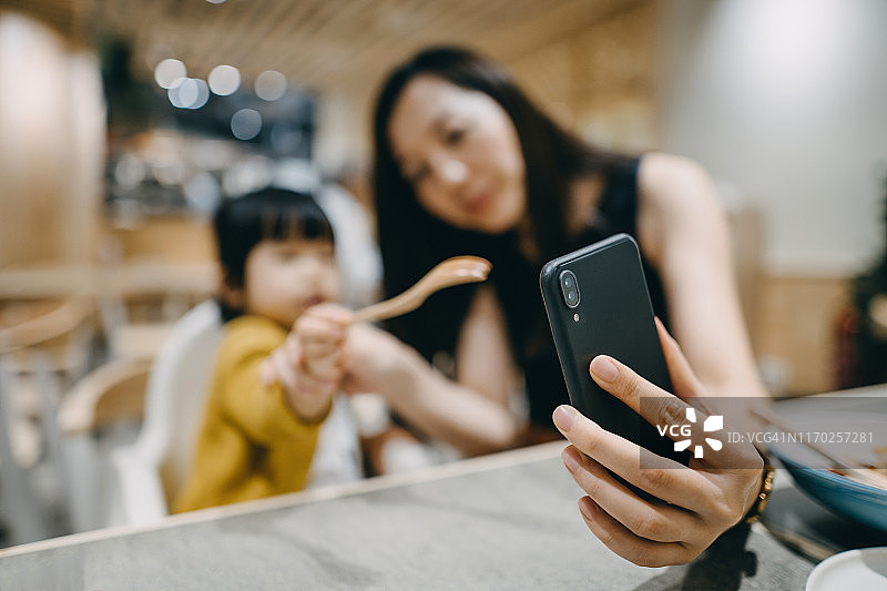 开心的年轻妈妈和可爱小女儿在餐厅用餐时手机视频通话招手图片素材