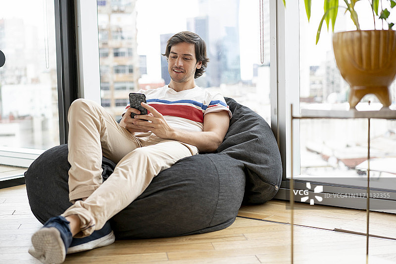 千禧一代男人在共享办公室内放松地坐在豆袋上，用智能手机聊天图片素材