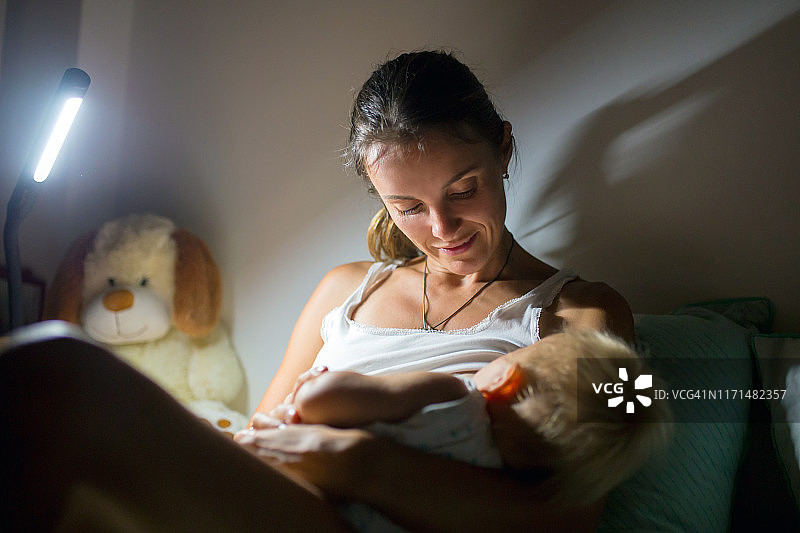 年轻美丽的母亲在昏暗灯光下母乳喂养新生儿。图片素材