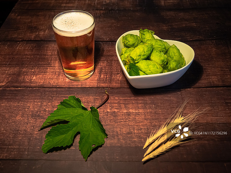 自酿啤酒：一杯白色泡沫的金色啤酒，心形瓷碗盛着啤酒花，啤酒花叶和大麦穗图片素材