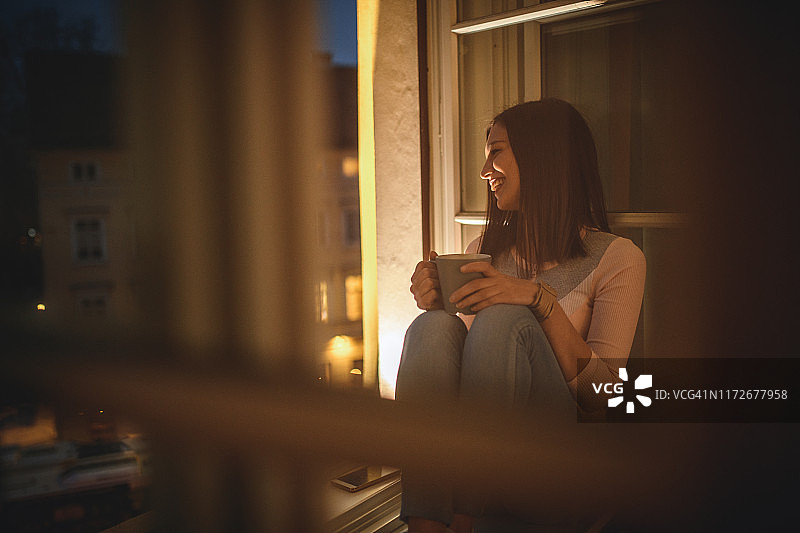 宁静的年轻女人在黄昏时分在开放的窗户边品茶图片素材