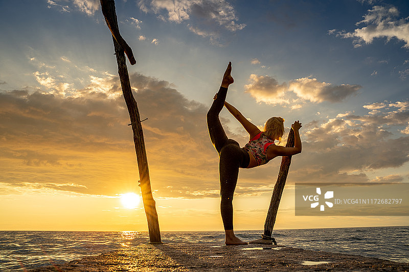 在日落时分海岸线上做瑜伽站立弓步姿势的年轻女人图片素材