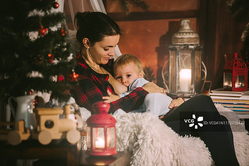 夜晚，母亲在圣诞树旁的舒适扶手椅上给幼儿儿子喂奶图片素材
