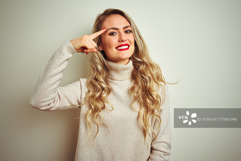 穿着高领毛衣的漂亮女人，在白色背景前微笑，指向头部，表示好主意图片素材