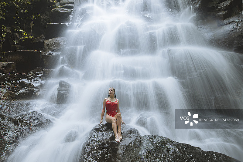 女人穿红色泳衣坐在印度尼西亚巴厘岛瀑布旁图片素材
