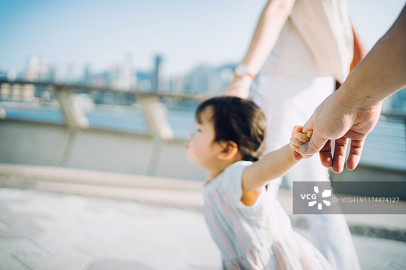 可爱的小女孩牵着父母的手，在阳光明媚的日子里漫步在城市长廊上图片素材