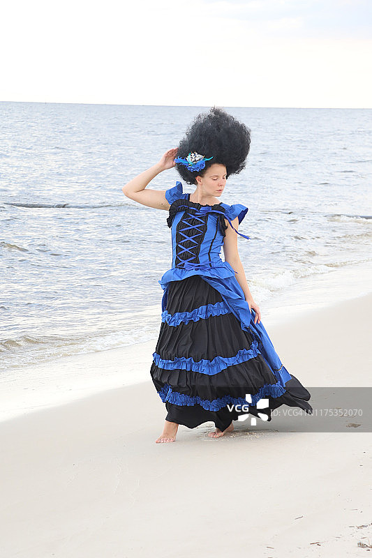 黑发美女身穿蓝黑色连衣裙站在海滩上，背景是海浪图片素材