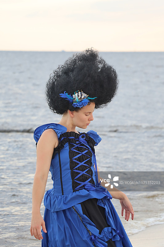 黑发美女身穿蓝黑色连衣裙站在海滩上，背景是海浪图片素材