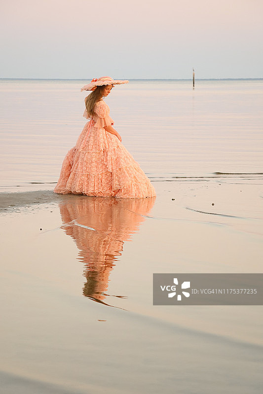 一位女士身穿桃色长裙，头戴相配的帽子，站在海滩上看着她的倒影图片素材