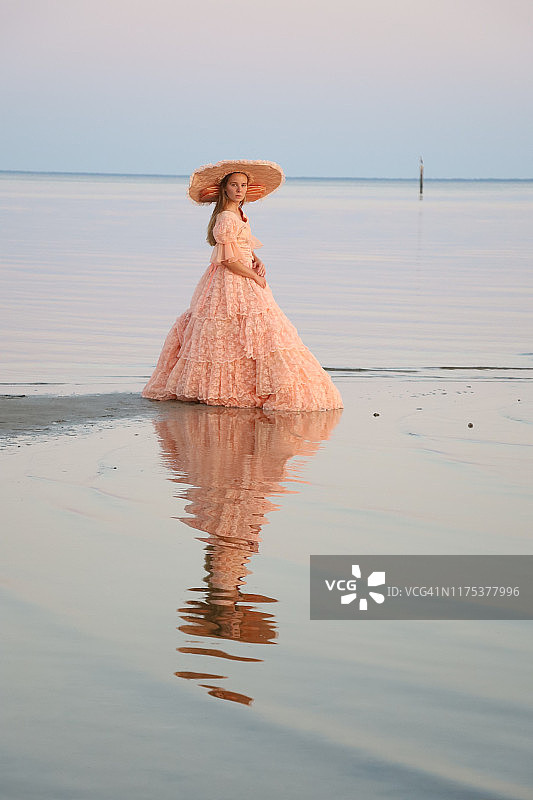 一位女士身穿桃色长裙，头戴相配的帽子站在海滩上，看着镜头图片素材