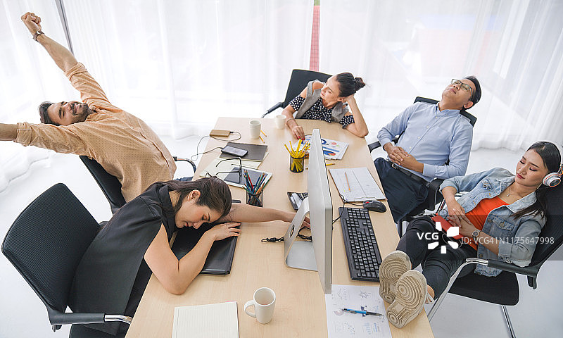 会议室里的咖啡休息时间：商务团队在办公桌旁休息。戴眼镜的年轻商人因工作过度而睡着，创意休闲男士在他的工作场所睡觉图片素材