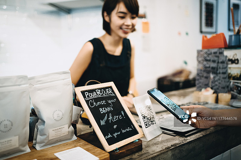 顾客扫描二维码，在咖啡馆里用智能手机进行快速便捷的非接触式支付，面前是一位面带微笑的咖啡师图片素材