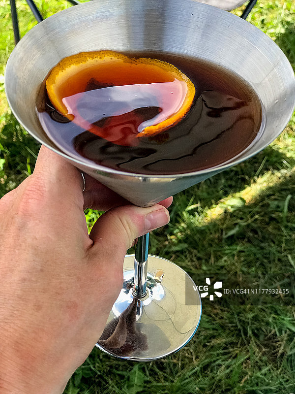 一杯冰爽的尼格罗尼鸡尾酒，由杜松子酒、味美思和金巴利混合而成，盛在金属马提尼杯中，背景是绿草图片素材