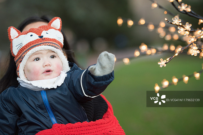亚洲宝宝戴着冬装、手套和狐狸帽子，在英国苏格兰爱丁堡王子街花园里伸手去拿圣诞灯饰图片素材