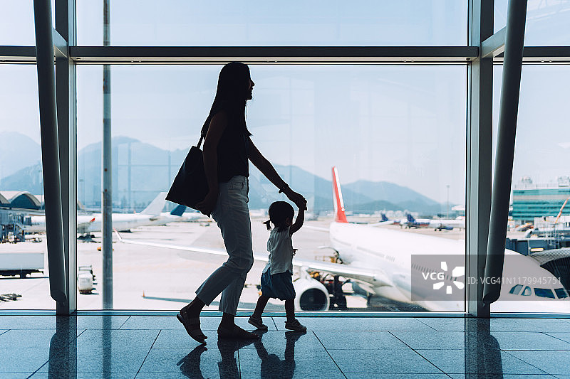 快乐的年轻亚洲母亲牵着可爱的女儿在机场候机时隔着窗户看飞机的剪影图片素材
