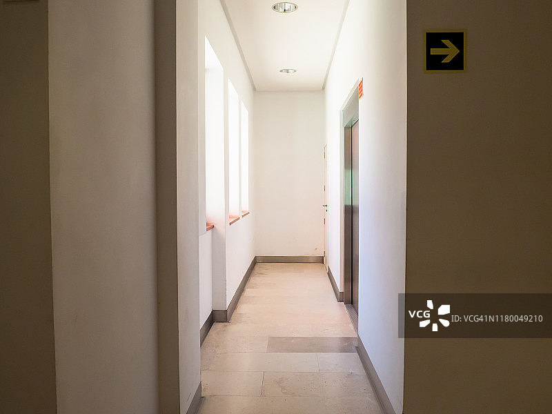 一个有窗户的白色走廊，自然光线射入，一个封闭的电梯和一个方向箭头图片素材