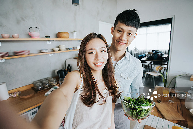 年轻的亚洲夫妇在厨房一起烹饪和准备食物时用智能手机自拍图片素材