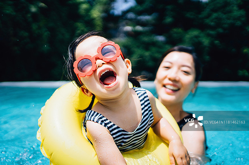 快乐的亚洲女童戴着太阳镜，笑容满面，与母亲在夏天的游泳池里享受家庭欢乐时光图片素材