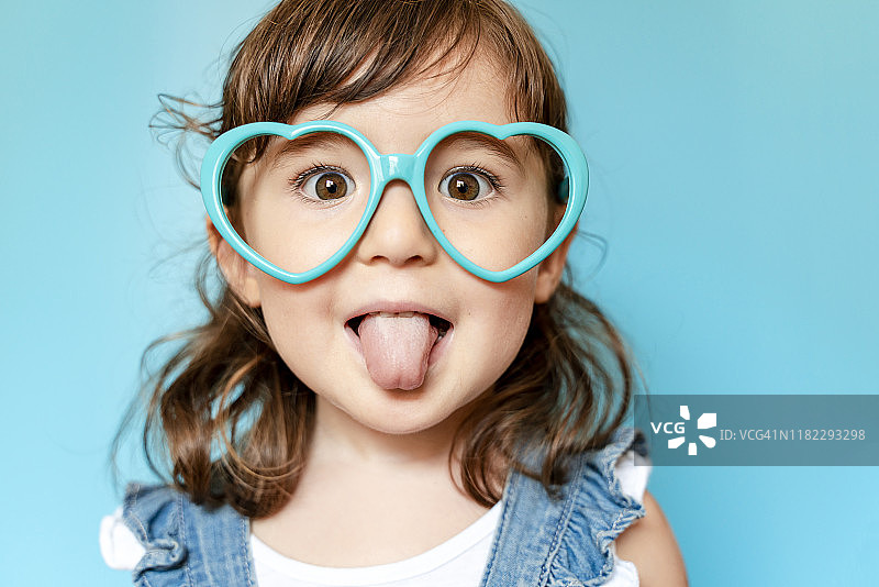 在蓝色背景下吐着舌头，戴着心形眼镜的可爱小女孩肖像图片素材