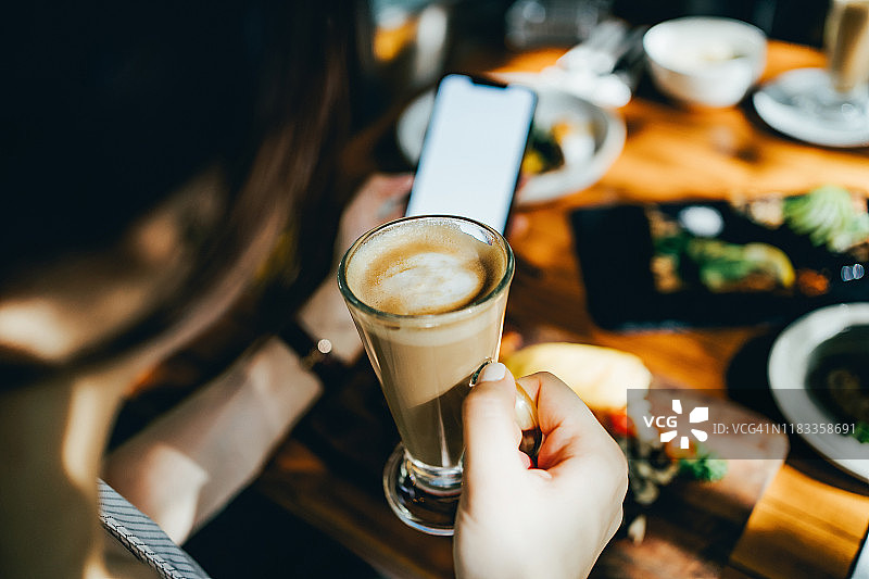 年轻女子在户外餐厅享用早午餐，沐浴在阳光下，喝咖啡和使用手机图片素材