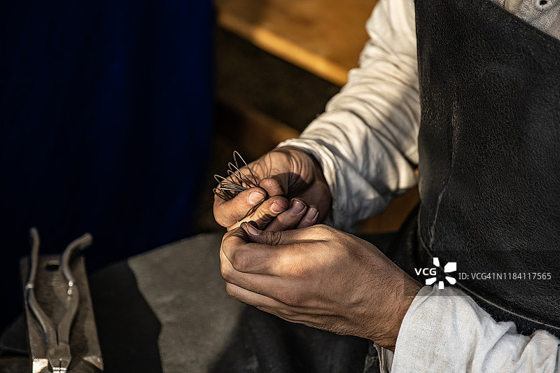 工匠的手。铁匠用手握住金属丝。制作锁子甲。古老的工艺。举办大师班。工作室。穿着旧的服装。附近有金属钳。工艺培训。图片素材