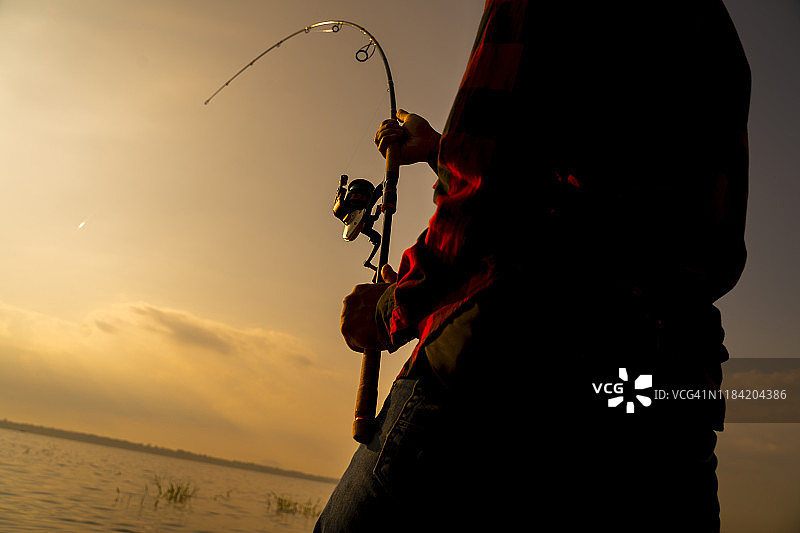 沉思的男人坐在水边，向右看。他手里拿着飞蝇竿。现在是傍晚，外面很冷。图片素材