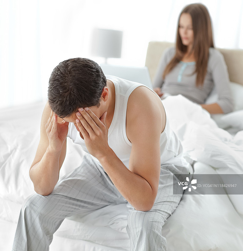 焦虑的男人坐在床上，女友在用笔记本电脑工作图片素材