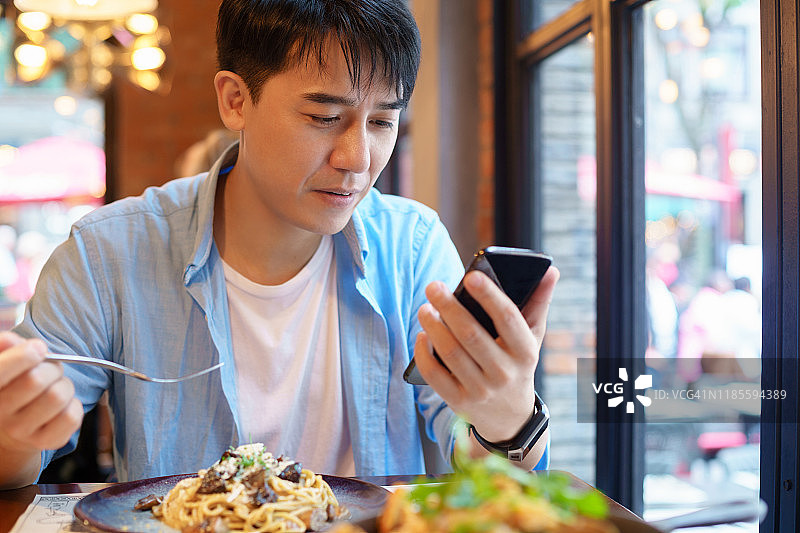 吃饭时使用智能手机的年轻亚洲男人图片素材