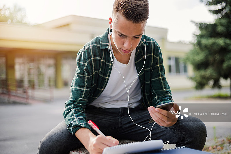 在校园里戴着耳机用智能手机听音乐写作的青少年图片素材