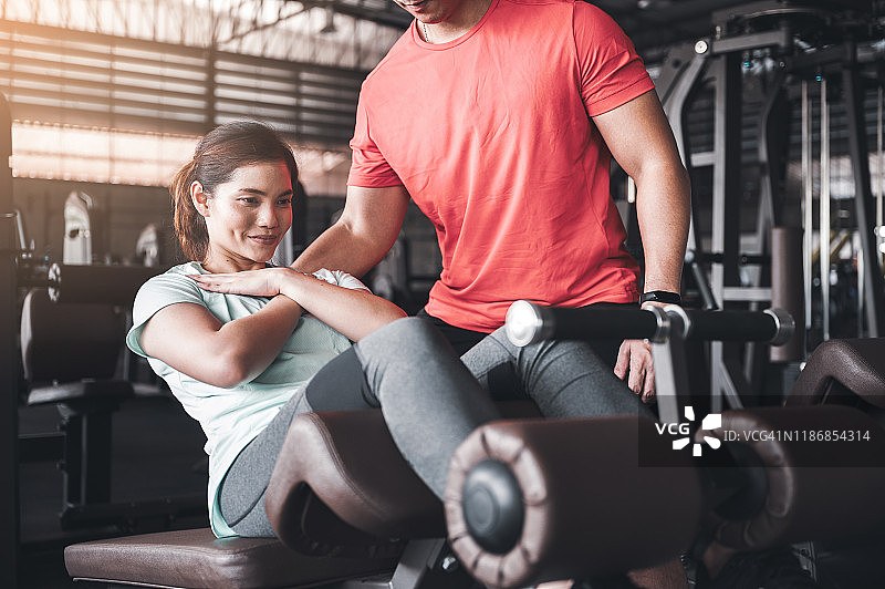 健身房概念。亚洲年轻女性在健身教练的指导下在健身房的仰卧起坐机上做仰卧起坐图片素材