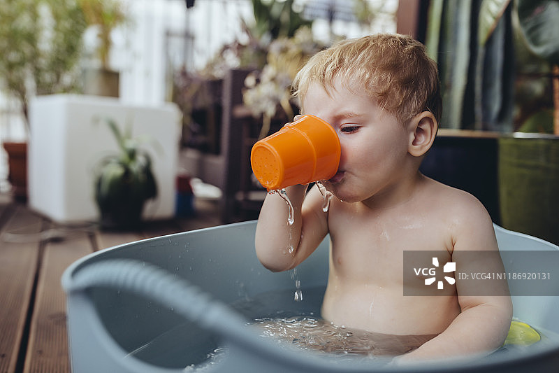 男婴坐在阳台的婴儿浴盆里喝水图片素材