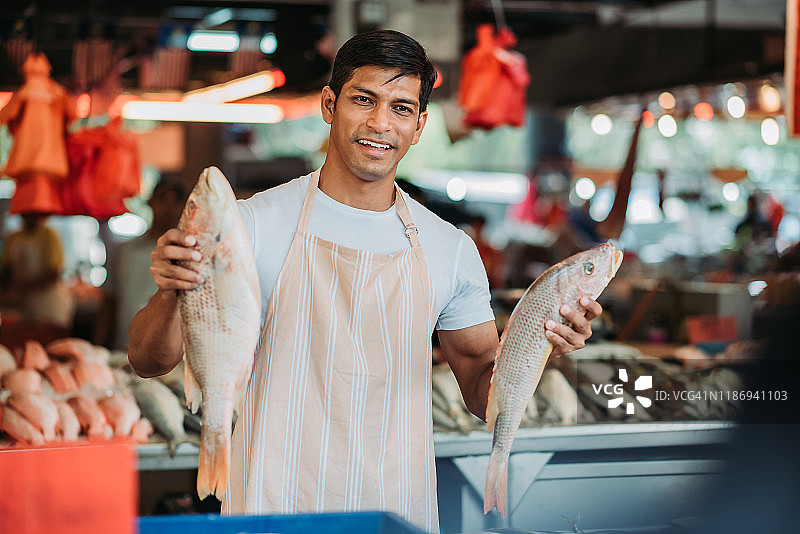 亚裔马来男鱼摊主手持鱼，面带微笑地看着顾客图片素材