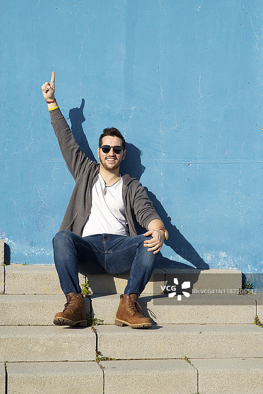 年轻络腮胡男子坐在户外楼梯上，抬手微笑远眺图片素材