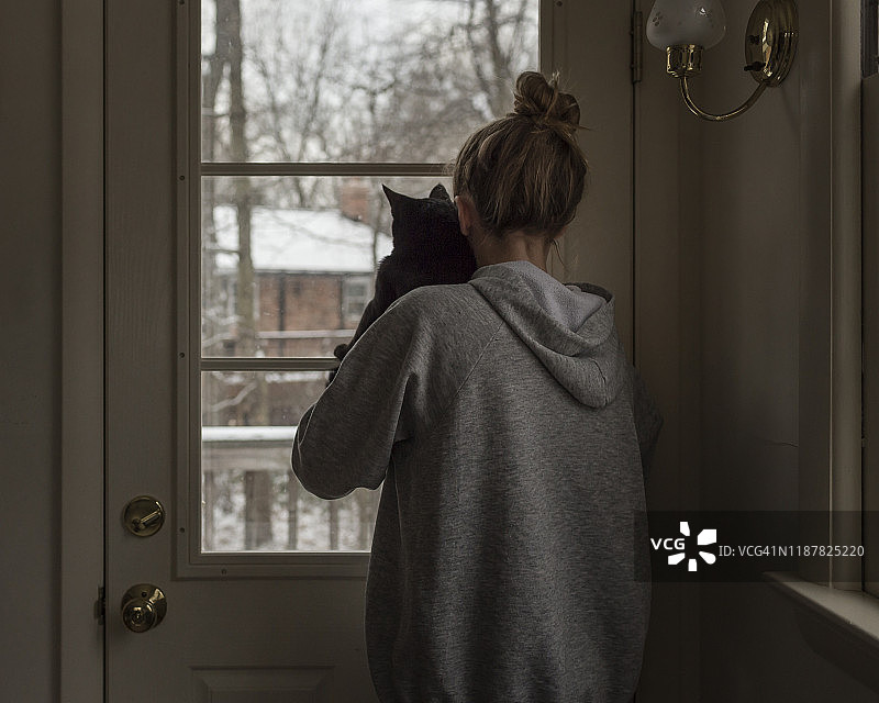 穿着连帽衫抱着猫在下雪天看窗外的少女图片素材