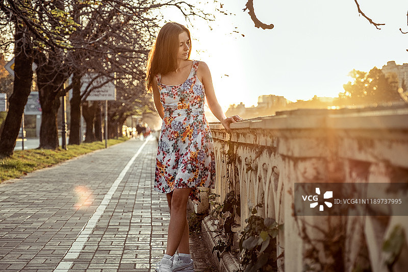 女孩走在人行道上。身穿浅色白色碎花夏季连衣裙。看着日落。河流。植物缠绕在栅栏上。图片素材