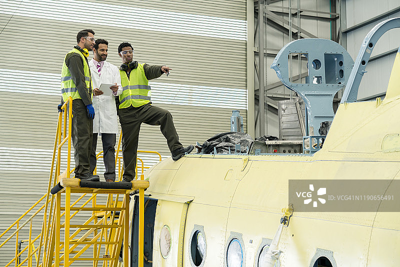 三名拉丁裔男子出现在黄色楼梯上，其中两名是机械师，一名是工程师，他们似乎在观看一架正在维修的黄色飞机图片素材